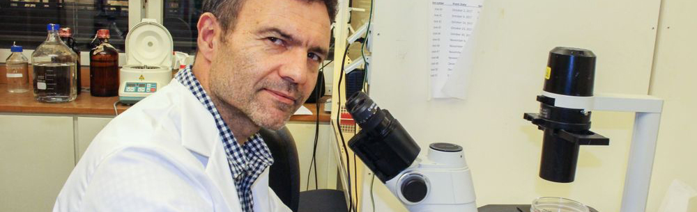 Forsker Fahri Saatcioglu sitter ved mikroskop og ser inn i kameraet. Foto.
